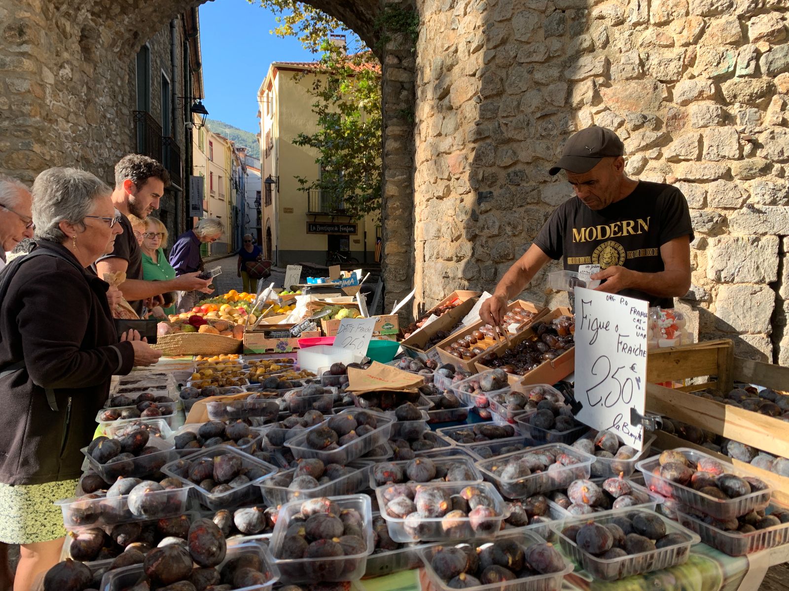 Étal d'un marché gascon sous les arcades d'une halle en pierre, un commerçant sert ses clients